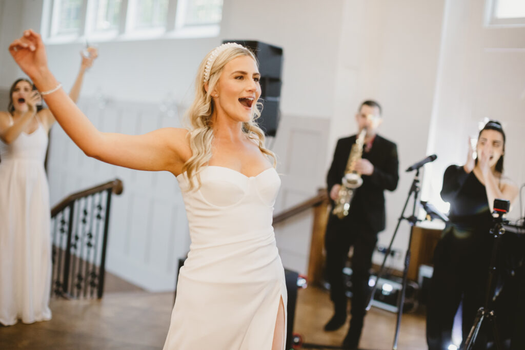 An image by Amy Lou Photography. A bride dances in the foreground with a saxophonist and singer playing in the background. Live vs. Recorded music